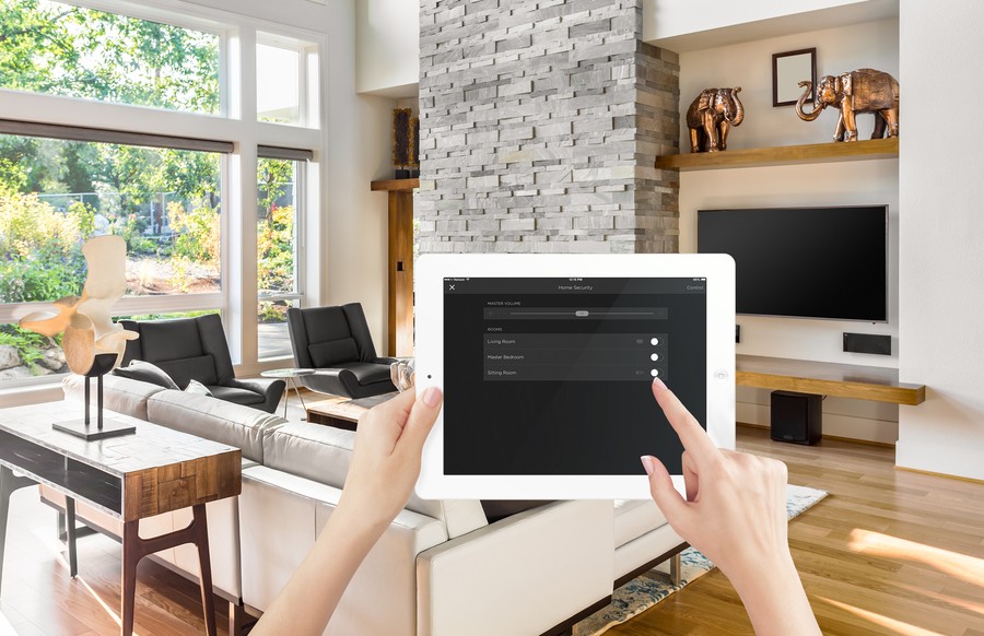 Hands holding a tablet with home automation controls on the screen. In the background is a bright, modern living room.  