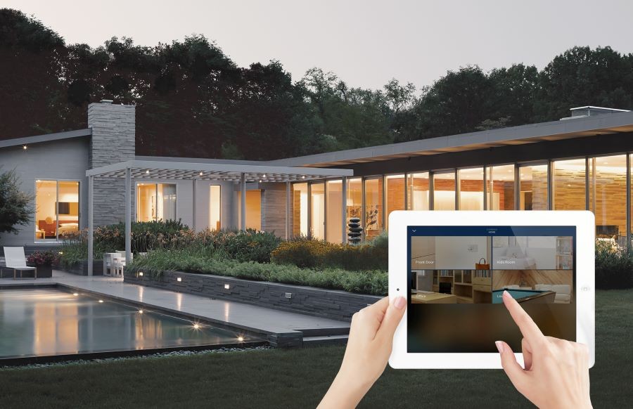 A backyard of a well-lit home with a pool. Overlayed hands are holding a tablet showing a Savant platform and security feed.