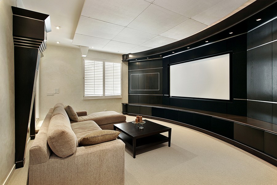 Tan, comfortable looking sofa and a dark wood, square coffee table set across from a dark, curved wall featuring a large television screen.