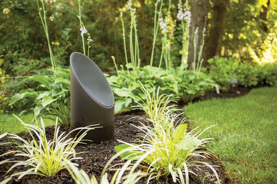 A sleek Coastal Source black outdoor speaker is discreetly placed among lush green plants and mulch in a landscaped garden, blending into the natural surroundings near a manicured lawn and trees.