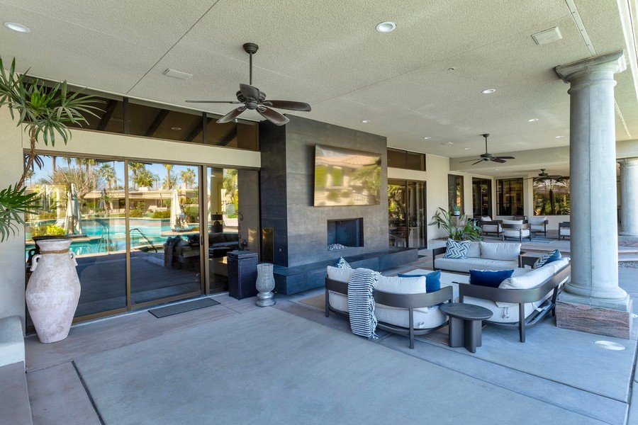 Luxury covered patio with modern outdoor seating, ceiling fans, fireplace, and mounted TV overlooking a pool and landscaped backyard.
