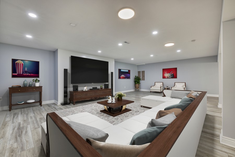 A modern home theater with a large TV, white sectional sofa, coffee table, artwork, and light gray walls with wood-look flooring.